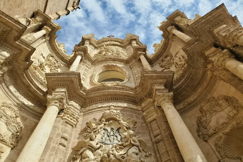 Vista de baixo para cima da fachada barroca ornamentada da Catedral de Valência, com detalhes em pedra, colunas e esculturas, destacando a rica arquitetura de Valência na Espanha.