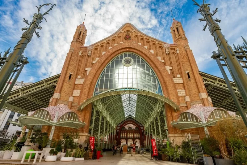 Fachada grandiosa do Mercado Central de Valência na Espanha, com sua arquitetura modernista em tijolos, vidro e ferro forjado, sob um céu azul e nuvens, um marco vibrante de Valência na Espanha.