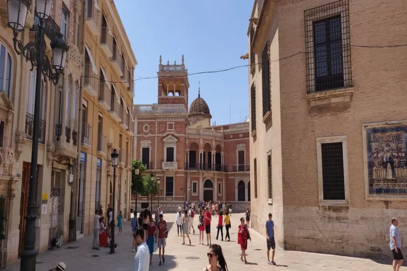 Praça movimentada em Valência na Espanha, com edifícios históricos de arquitetura clássica, incluindo o Palácio do Marquês de Dos Águas, sob um céu azul, representando a beleza de Valência na Espanha.