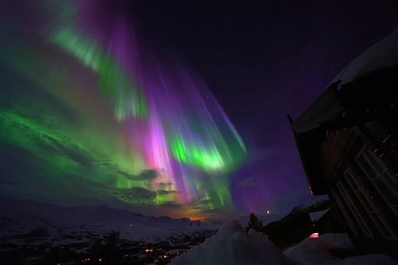 Aurora boreal em tons de verde e roxo iluminando o céu noturno sobre neve e cabana rústica na Noruega