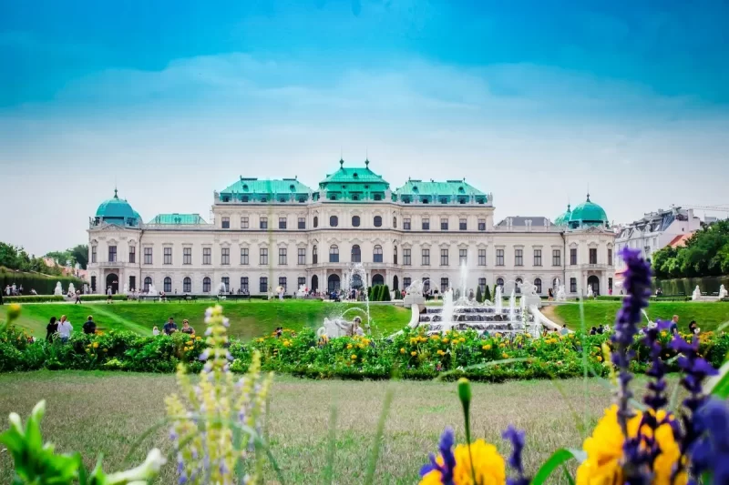O Palácio Belvedere em Viena, capital da Áustria, com jardins, flores coloridas e uma fonte em primeiro plano.