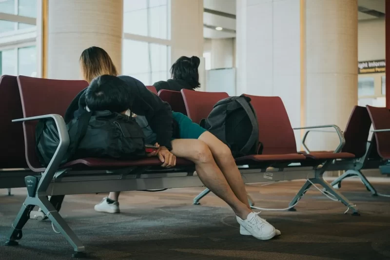 Pessoa dormindo curvada sobre uma mochila em assentos de aeroporto, com os pés no banco, representando o cansaço e os efeitos do jet lag.