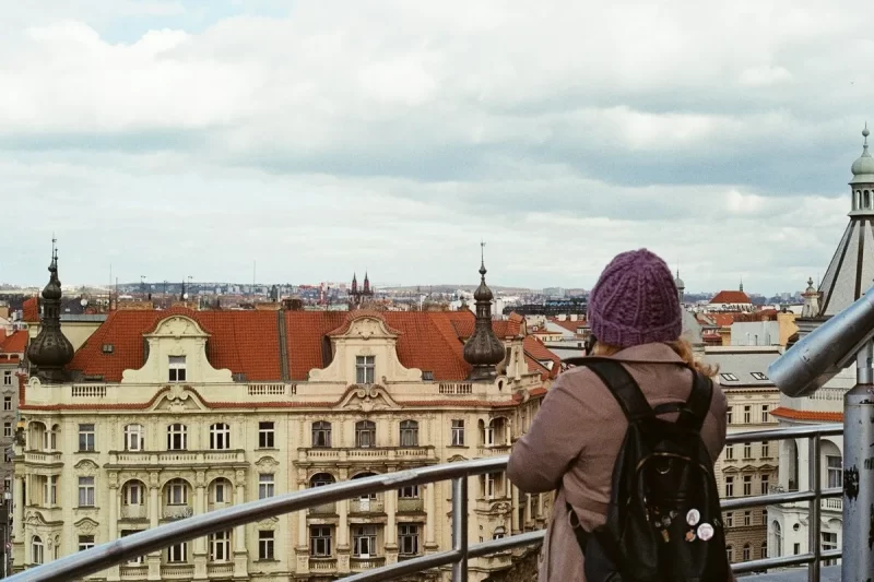 Uma pessoa de costas, usando gorro roxo e mochila preta, observando a paisagem urbana de uma cidade europeia com telhados de terracota e arquitetura clássica, de um ponto de vista elevado.