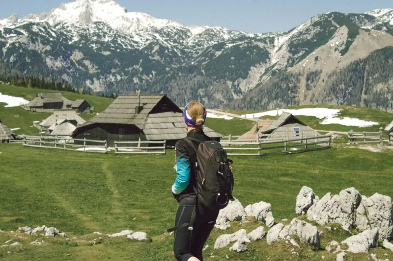 Uma pessoa de costas, com mochila, observando um vale alpino com cabanas de madeira tradicionais e montanhas nevadas ao fundo, exemplificando um mochilão na Europa.