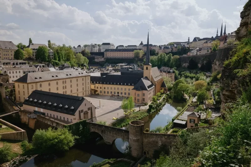 Vista panorâmica da cidade de Luxemburgo, capital do país mais rico da Europa, com prédios históricos, rio, ponte e vegetação exuberante.