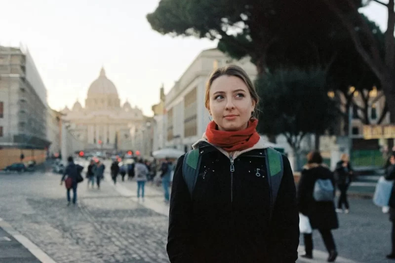 Mulher jovem com mochila e cachecol vermelho, olhando para o lado em uma rua europeia, com a Basílica de São Pedro ao fundo, simbolizando a importância do seguro viagem na Europa.