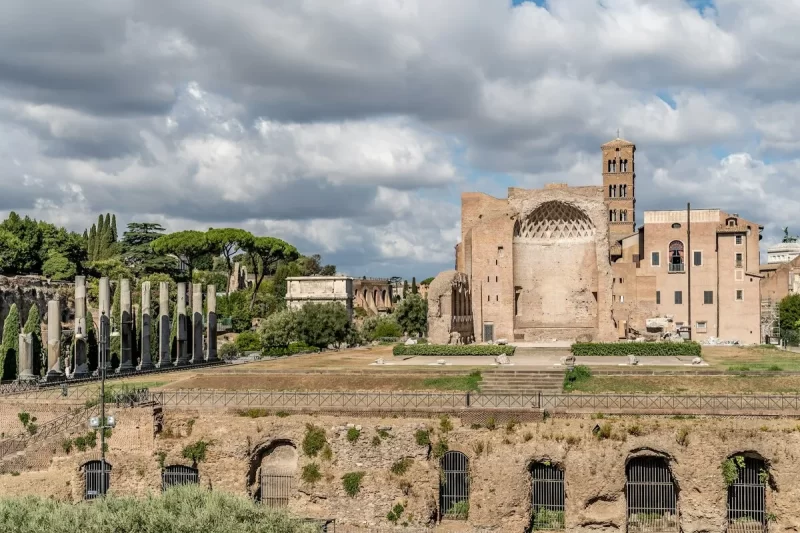 Vista ampla das ruínas do Fórum Romano em Roma, com colunas, edifícios antigos de tijolos e vegetação, sob um céu parcialmente nublado, representando um complexo de sítios arqueológicos.
