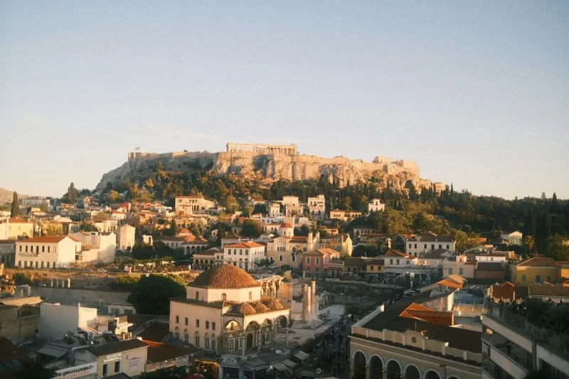 Paisagem panorâmica da cidade de Atenas ao entardecer, com a Acrópole iluminada no topo da colina, vista de um bairro histórico.