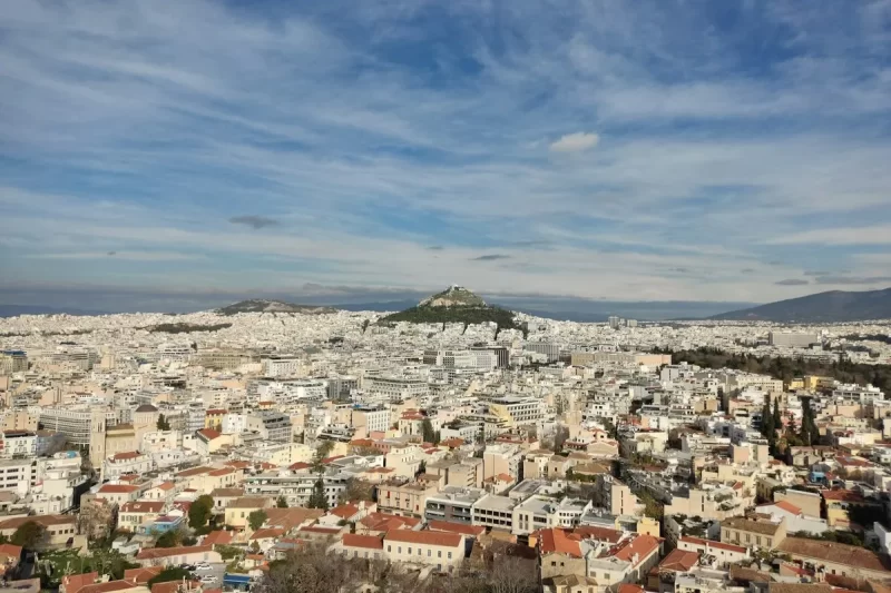 Vista panorâmica aérea de Atenas, com a densa área urbana, o Monte Licabeto e o Monte Filopapo em destaque, sob um céu azul com nuvens brancas.