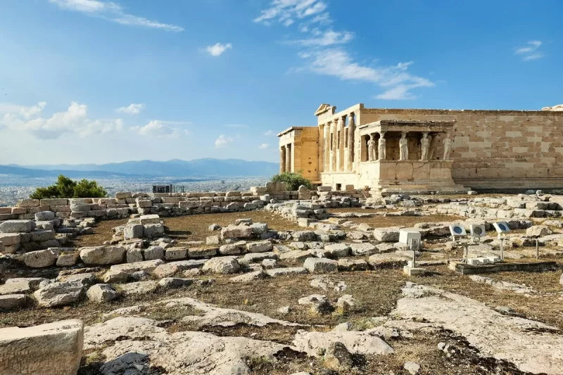Ruínas históricas da Acrópole de Atenas com o Erecteion em destaque e as Cariátides, sob um céu azul, com vista panorâmica da cidade.