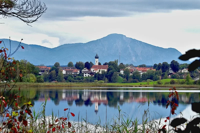 Vilarejo com igreja na margem de um lago, com montanhas ao fundo, representando a paisagem serena da Baviera.