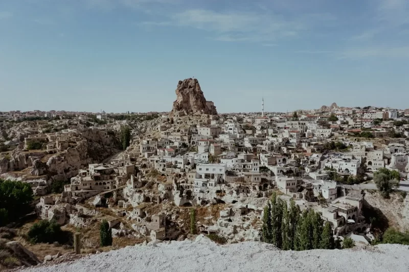 Vista panorâmica da cidade de Göreme na Capadócia, Turquia, mostrando casas e hotéis de pedra esculpidos nas encostas de montanhas e ao redor de uma grande formação rochosa icônica. A paisagem é árida com céu azul claro.