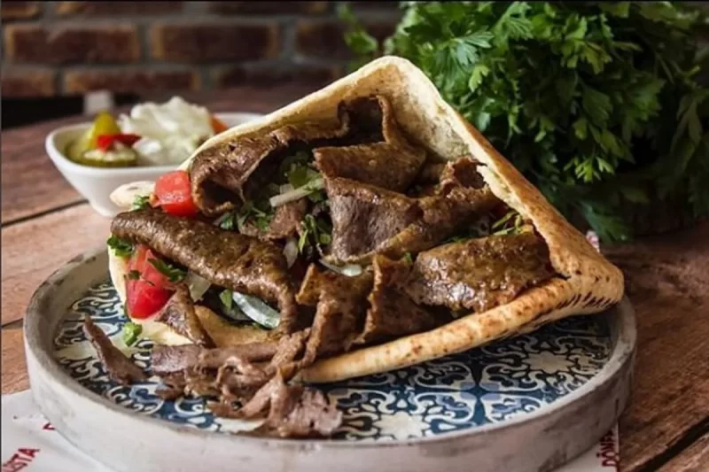 Um Kebab ou Gyro farto e apetitoso, recheado com carne grelhada, tomate, cebola e salsinha, servido em um prato com estampa e acompanhado de picles e salada. A imagem destaca a culinária de rua de Chipre.