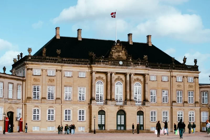 Fachada do Palácio de Amalienborg em Copenhague, um majestoso edifício histórico com a bandeira dinamarquesa hasteada, com pessoas visitando a praça em frente.