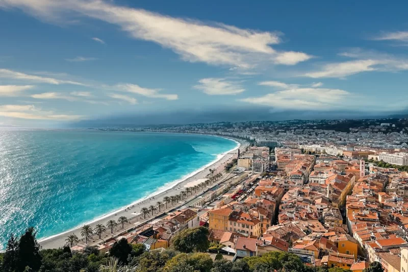 Vista aérea da orla de Nice, França, com o mar azul, a praia e a Promenade des Anglais, e a cidade com telhados laranjas ao fundo.