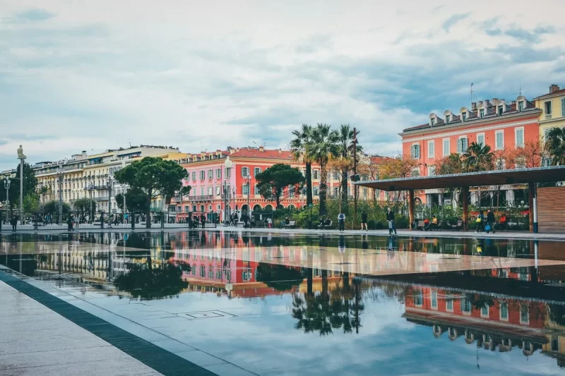 Reflexo dos prédios de cores vivas e palmeiras no chão espelhado da Praça Masséna em Nice, França, em um dia nublado.