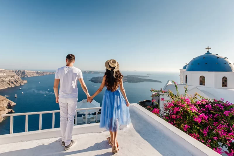 Um casal de mãos dadas, visto de costas, em um terraço em Santorini, com o mar azul-claro, ilhas e a arquitetura branca e cúpulas azuis ao fundo. A imagem representa o romance e a beleza de Santorini na Grécia.
