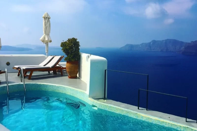 Uma piscina infinita de cor azul-clara em um terraço, com uma vista panorâmica para o mar azul-escuro e ilhas ao longe, em Santorini, na Grécia.