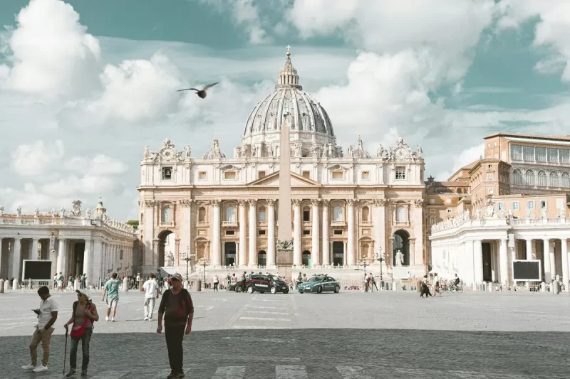 Vista frontal da monumental Basílica de São Pedro e da Praça de São Pedro, o coração da Cidade do Vaticano. A imagem mostra a grandiosa cúpula e a fachada, com turistas a passear no chão de paralelepípedos.