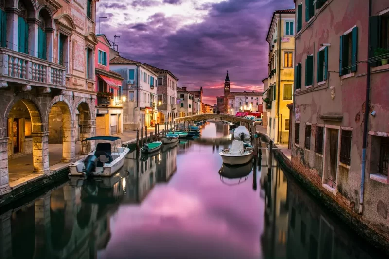 Veneza na Itália ao pôr do sol, com canal estreito, barcos e reflexo roxo.