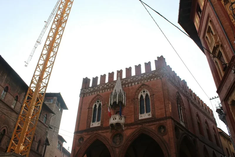 Vista de um edifício histórico em Bolonha, Itália, com uma torre medieval, uma bandeira da Itália e um guindaste de construção ao fundo.