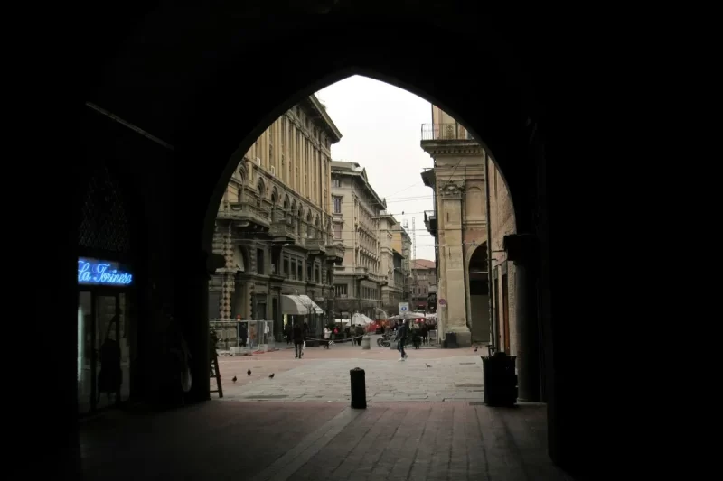 Vista através de um arco de uma rua estreita e histórica em Bolonha, Itália, com edifícios de arquitetura medieval e pessoas caminhando.