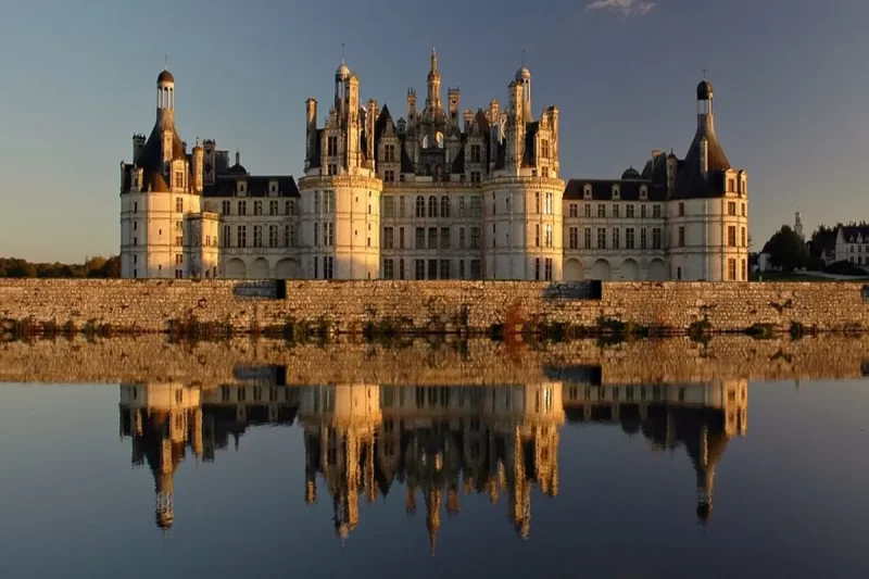 O Castelo de Chambord, um castelo medieval de cor clara, refletido na água de um lago. A imagem representa a beleza e a grandiosidade de castelos medievais.