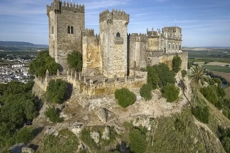 Um castelo medieval de pedra, com várias torres e ameias, situado no topo de uma colina, em um dia ensolarado. A imagem mostra a grandiosa arquitetura dos castelos medievais.