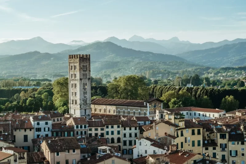 Uma vista panorâmica da cidade de Lucca, uma das mais encantadoras cidades na Toscana, com a icónica Torre Guinigi a sobressair dos telhados de terracota. Ao fundo, as montanhas e a paisagem verde da região.