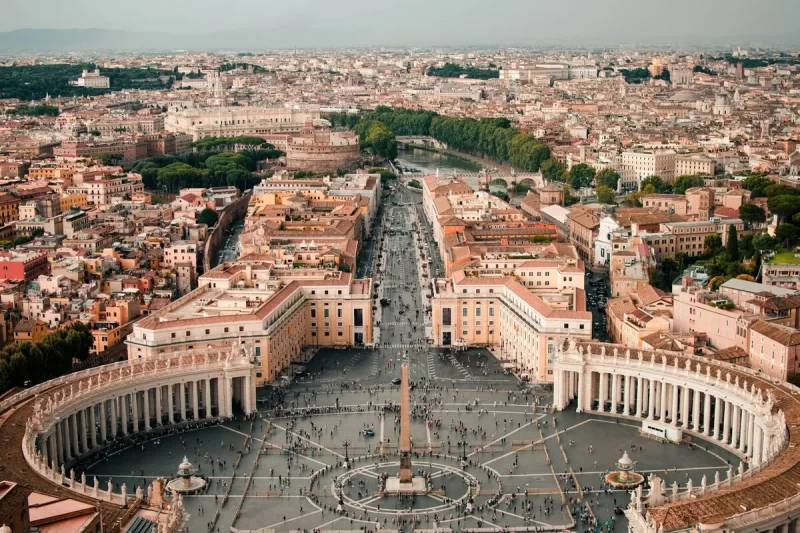 Vista aérea da Praça de São Pedro e da Via della Conciliazione, mostrando o Vaticano e o Castel Sant'Angelo, com a vasta cidade de Roma ao fundo. A imagem capta a grandiosidade dos monumentos e a extensão da cidade.