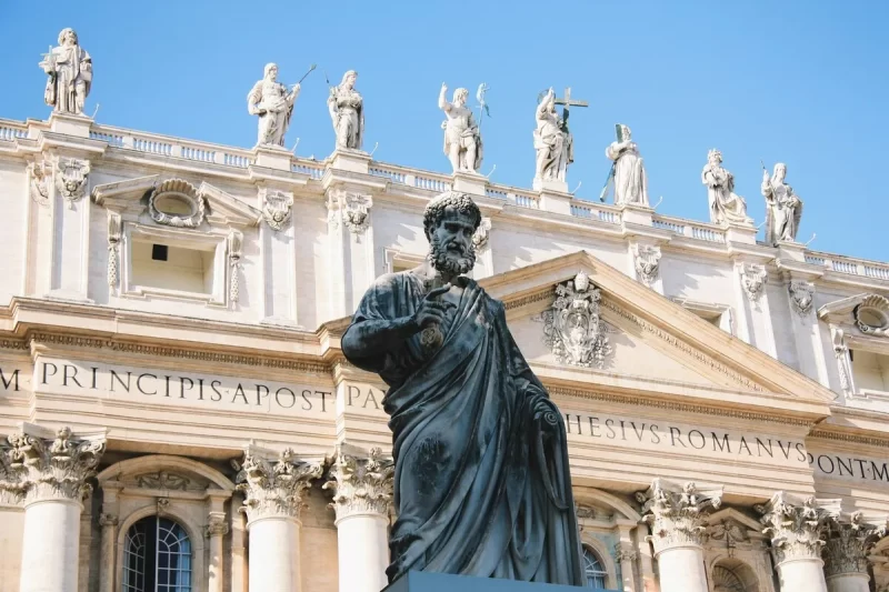 A famosa estátua de bronze de São Pedro, o guardião das chaves, em frente à fachada da Basílica de São Pedro, no Vaticano. A imagem foca no simbolismo religioso e na arquitetura detalhada do local.