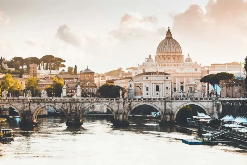 Vista da Basílica de São Pedro no Vaticano, refletida no Rio Tibre, com uma ponte de pedra em primeiro plano. A imagem capta o icónico monumento contra o céu dourado, numa paisagem que mistura arquitetura e natureza.