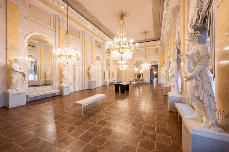 Sala elegante de palácio histórico com piso de madeira, grandes lustres de cristal, esculturas clássicas de mármore e paredes decoradas em tons de dourado e bege.