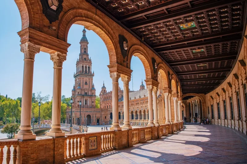 Vista da Plaza de España em Sevilha através de arcos semicirculares, mostrando torre, galerias e arquitetura renascentista-mourisca.