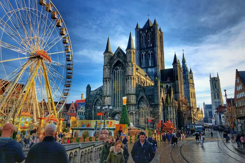 Vista da Catedral de São Nicolau em Ghent com uma roda-gigante e feira ao redor, além de turistas caminhando pela Korenmarkt.