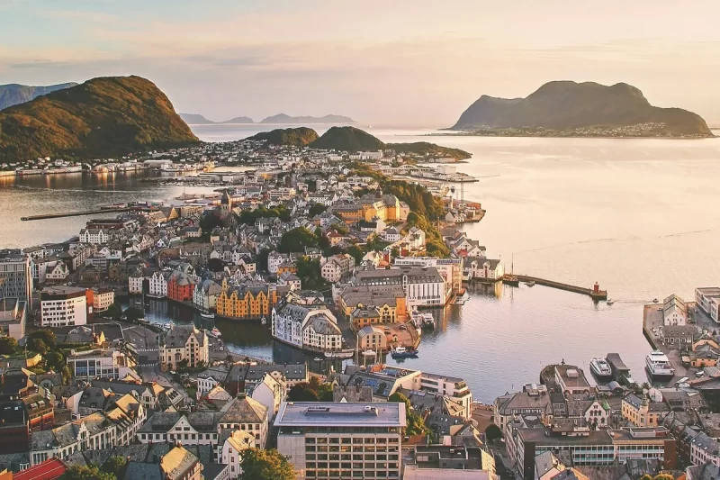 Imagem aérea de Ålesund, na Noruega, com casas coloridas em estilo Art Nouveau cercadas por fiordes e montanhas ao entardecer.